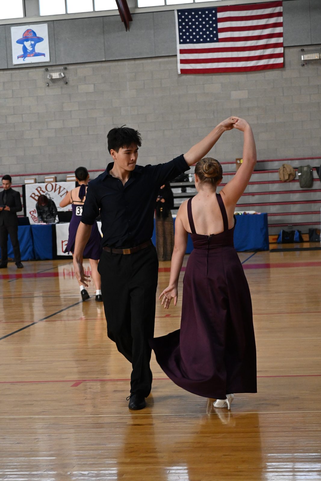 A couple in an underarm turn on a competition floor, other couples behind them.